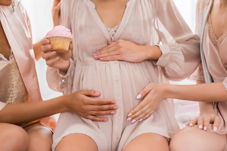 Two friends touch the belly of a pregnant woman holding a cupcake