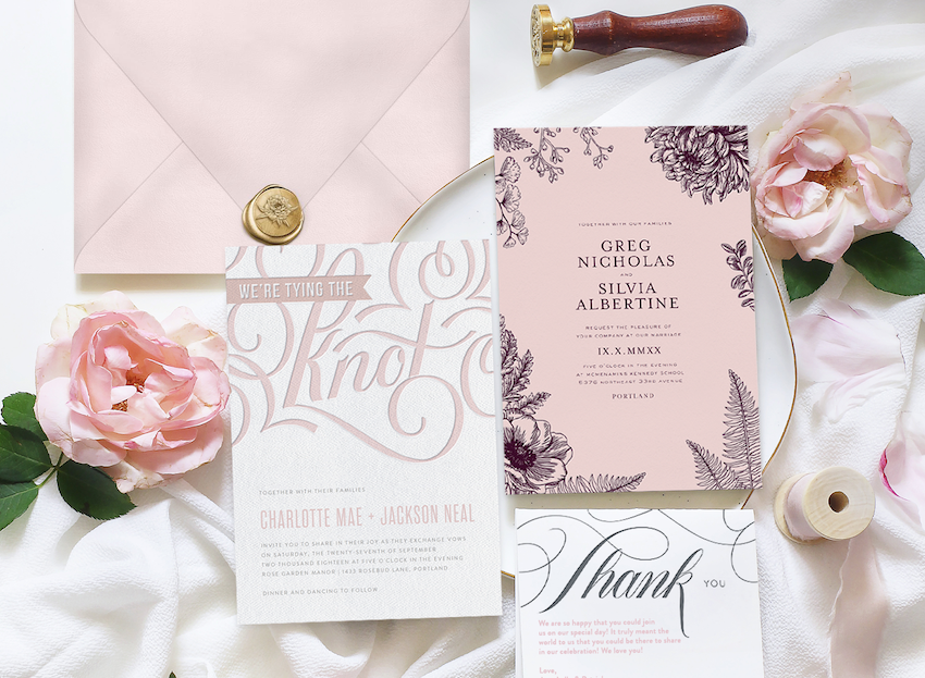 Three letterpress wedding invitations laid out on fabric with flowers, ribbons, and a wax seal