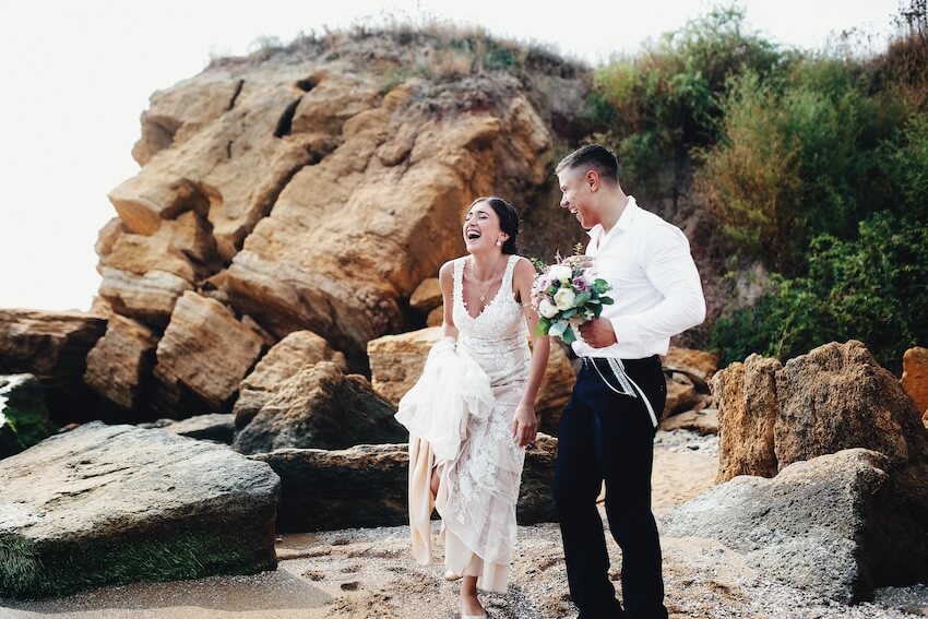 Newlywed questions: bride and groom walking at a beach with boulders in the background