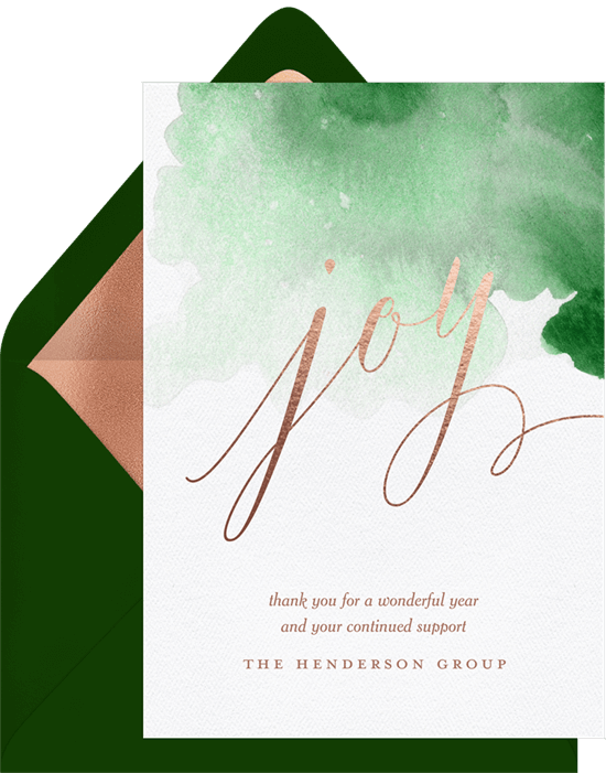 A holiday card featuring a handwritten script reading "joy" in a rose gold foil. Behind the word "joy" there is a splash of green watercolor.