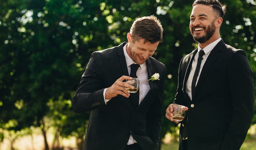 Wedding party roles: groom and his best man laughing while holding their glasses