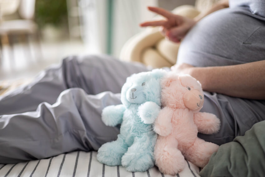 Twin pregnancy announcement: pregnant woman sitting on a bed beside two teddy bears