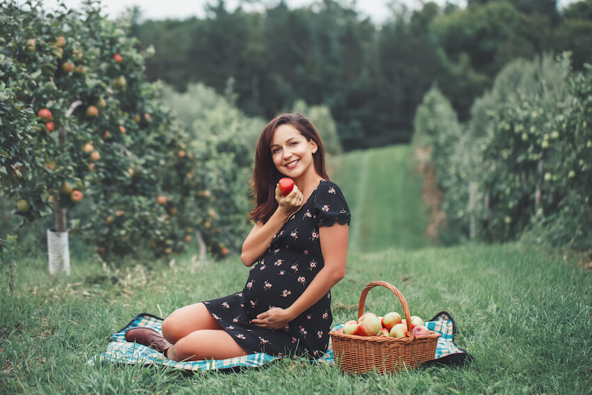 Fall pregnancy announcement: pregnant woman smiling at the camera while holding an apple